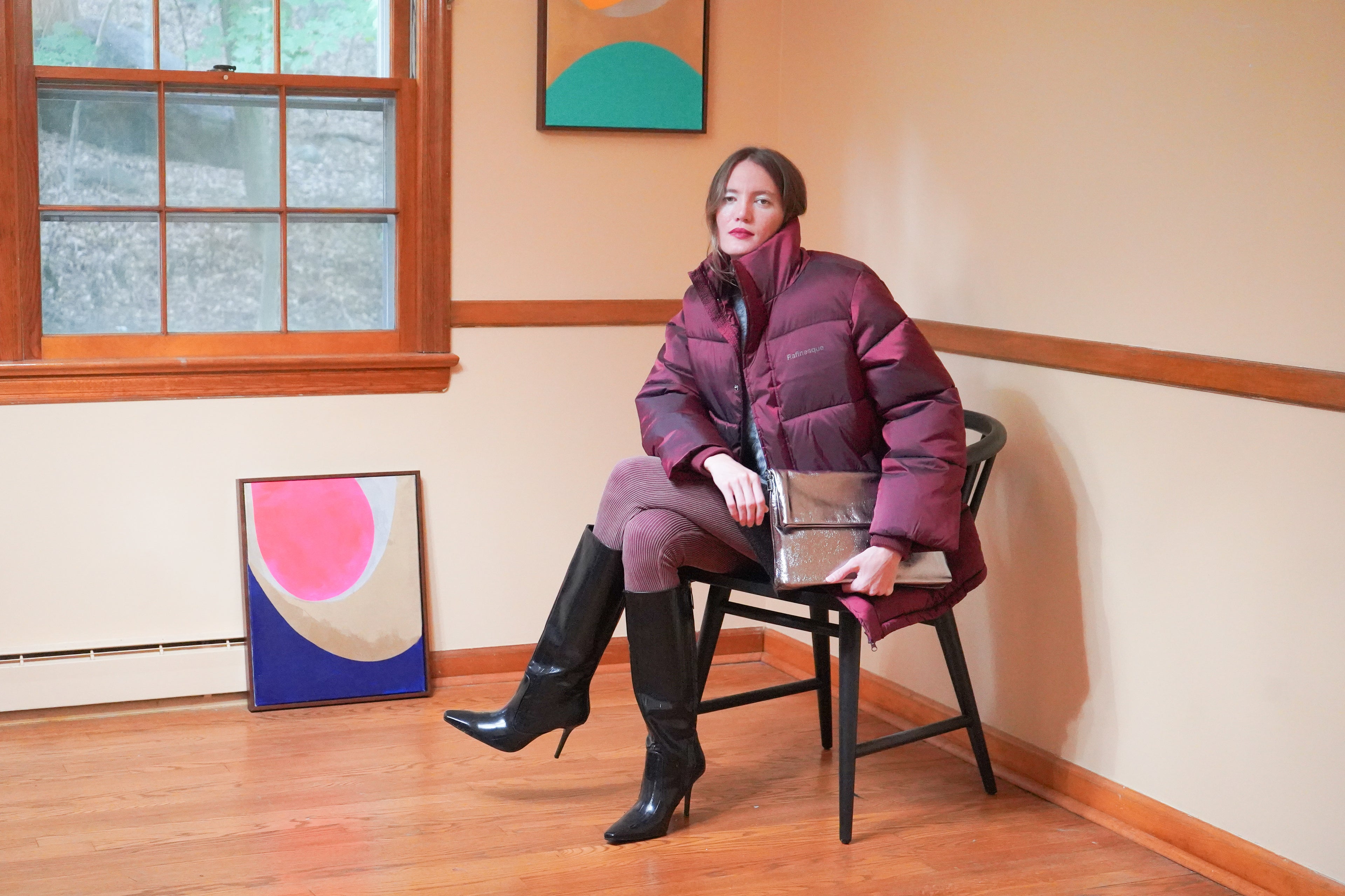Dark plum puffer jacket, matching plum pants, black knee-high boots, and holding a metallic silver Evelyn clutch.